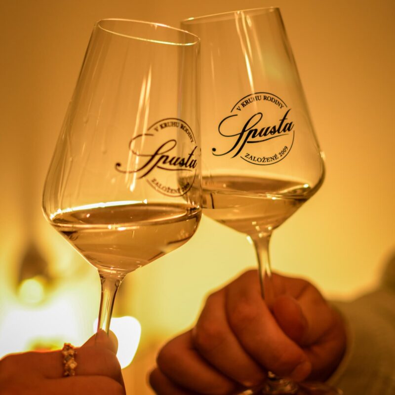 Close-up of elegant wine glasses with the logo 'Spusta' engraved, softly lit in a warm ambiance, with a blurred figure in the background.