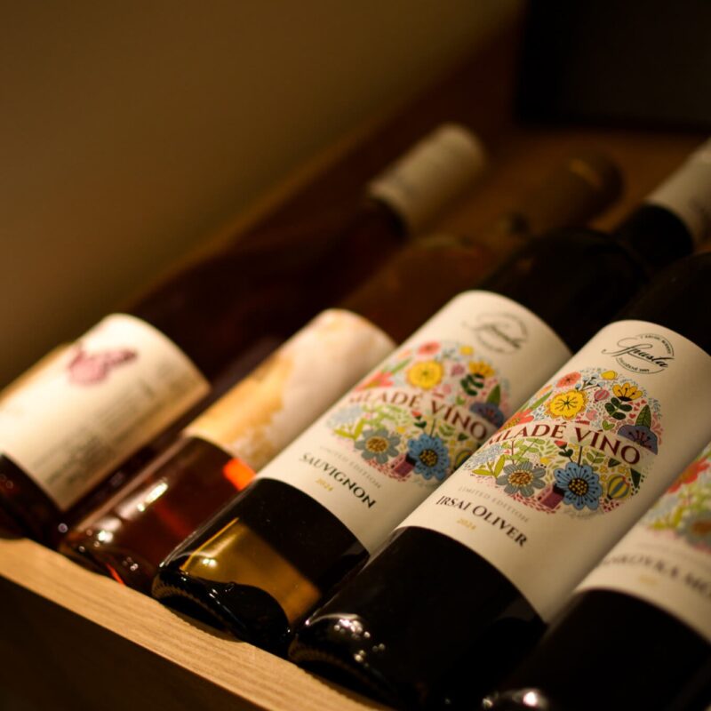 An assortment of Slovak wines displayed on a wooden shelf, featuring colorful floral labels on bottles of 'Mladé Vino' by Spusta, highlighted in soft, warm lighting