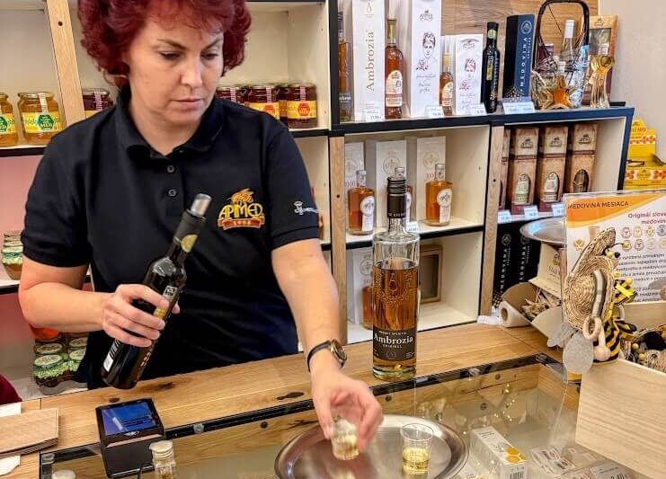 A shopkeeper at Apimed Honey Shop in Bratislava, pouring a sample of mead for a tasting, surrounded by shelves filled with honey and mead products.