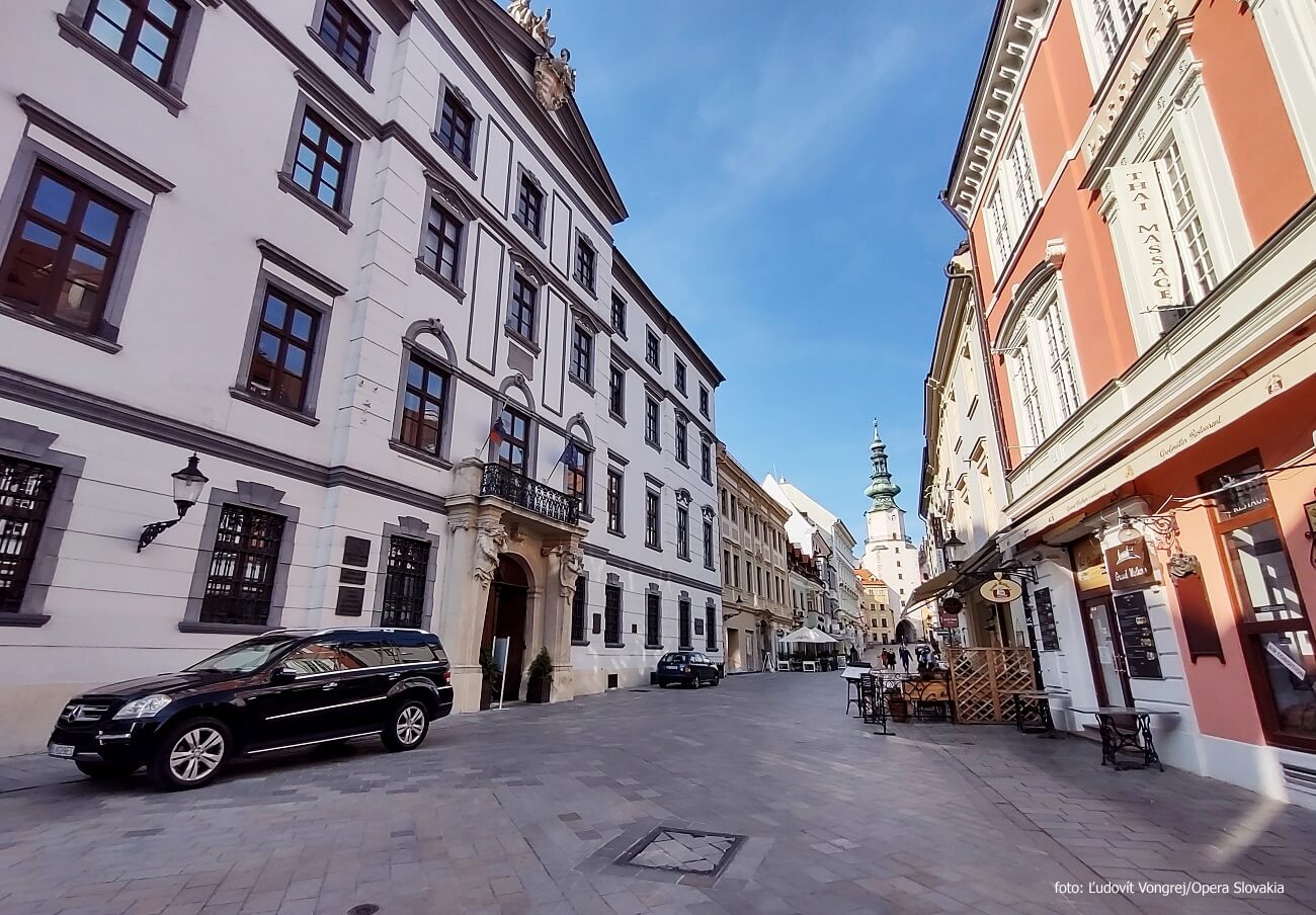 A view of Michalska Street in Bratislava featuring the Hungarian Royal Chamber Palace on the left. The palace's elegant Renaissance facade stands out, while other historic buildings and shops line the street. St. Michael's Gate is visible in the distance under a clear blue sky.