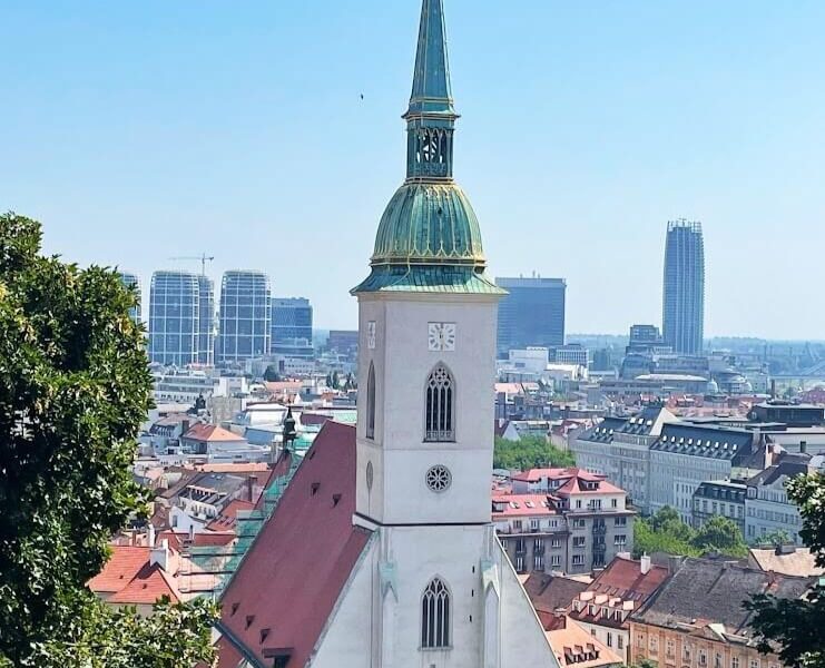 Aerial view of St. Martin's Cathedral in Bratislava with its tall green and gold spire standing prominently among the city's modern skyline and historical red rooftops.