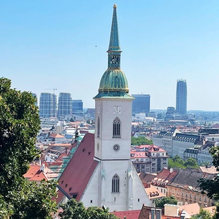 Aerial view of St. Martin's Cathedral in Bratislava with its tall green and gold spire standing prominently among the city's modern skyline and historical red rooftops.