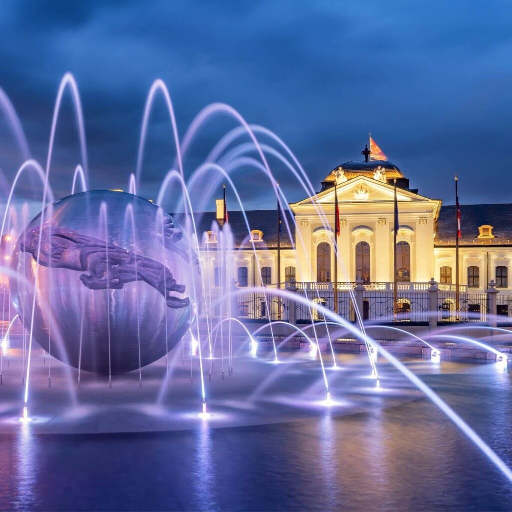 Illuminated Grassalkovich Palace in Bratislava at night, with a stunning fountain and modern sculpture in the foreground