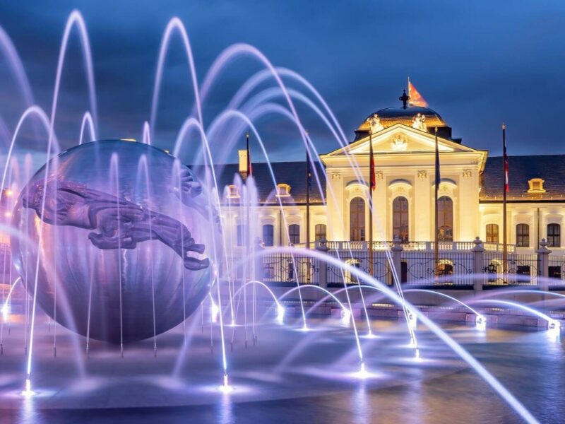 Illuminated Grassalkovich Palace in Bratislava at night, with a stunning fountain and modern sculpture in the foreground