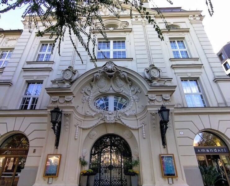 Front view of the Reduta Building in Bratislava, showcasing its ornate Baroque-style architecture with intricate sculptures and an arched entrance framed by two lanterns. The cream-colored facade reflects the historic significance of this cultural venue.