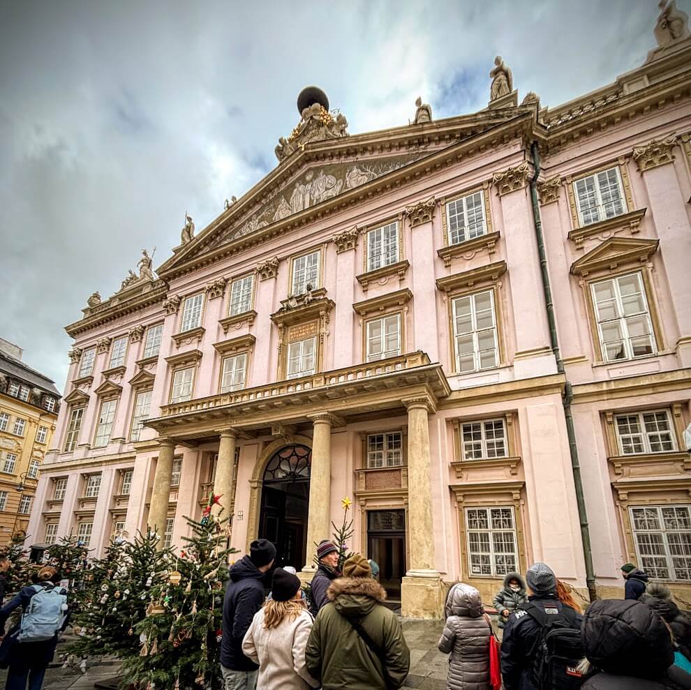 Primacial Palace, a grand pink neoclassical building with ornate architectural details, Corinthian columns, and statues on the roof. The scene is festive, with Christmas trees and a group of people gathered in front, suggesting a winter holiday settin