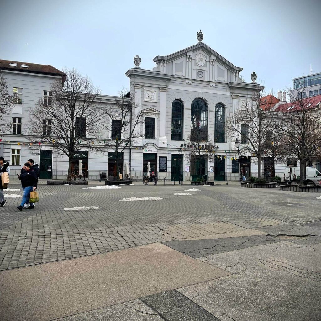 Present-day view of the Bratislava Old Town Market Hall, a white historic building in the city center surrounded by a cobblestone square.