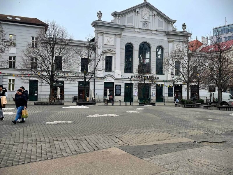 Present-day view of the Bratislava Old Town Market Hall, a white historic building in the city center surrounded by a cobblestone square.