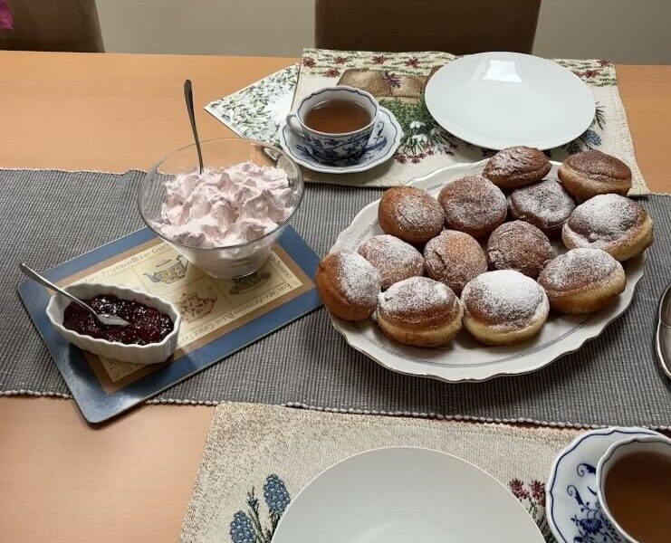 A cozy table setting with a plate of freshly made Šišky, traditional Slovakian donuts, dusted with powdered sugar. Accompanying them are a bowl of whipped fruit cream, a dish of jam, and cups of tea, ready for a delightful homemade trea