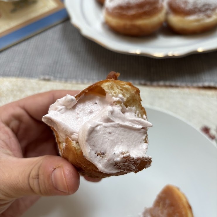 A close-up of a Slovakian šiška, showing its fluffy, golden-brown texture topped with a generous spread of whipped fruit cream. In the background, more powdered sugar-dusted donuts sit on a plate, inviting a sweet indulgence.