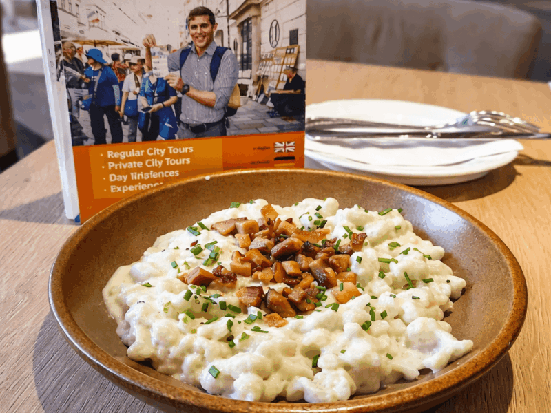 A bowl of bryndzové halušky — traditional Slovak potato dumplings smothered in creamy bryndza sheep cheese, topped with crispy bacon bits and fresh chives — served in a rustic brown ceramic bowl at a Bratislava restaurant, with a Bratislava City Tours brochure in the background.