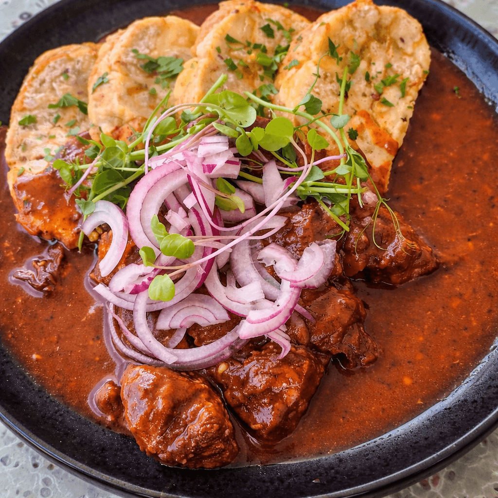 A generous serving of Slovak beef goulash — rich, paprika-spiced braised beef in a deep brown sauce — plated in a dark ceramic bowl and topped with sliced raw red onion and fresh microgreens, served alongside golden pan-fried bread dumplings (žemľovka/knedľa) garnished with herbs, at a Bratislava restaurant.