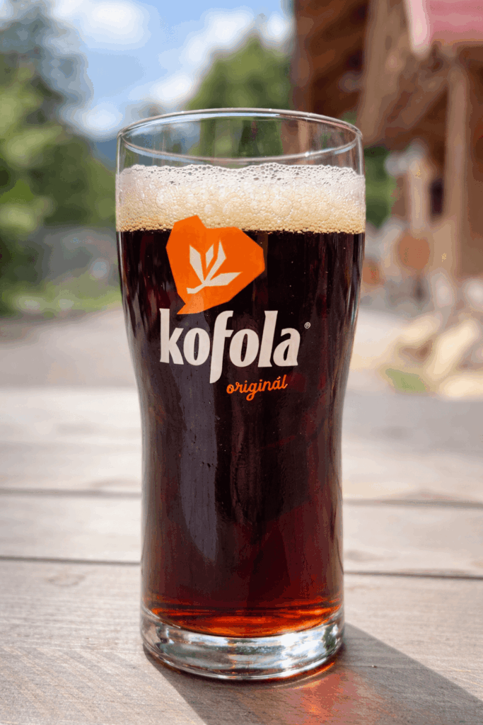 A tall glass of Kofola Originál — the iconic Slovak herbal cola — with a frothy head, served at an outdoor terrace with a mountain forest backdrop on a sunny summer day. Kofola is a beloved Central European soft drink and a must-try when visiting Slovakia.