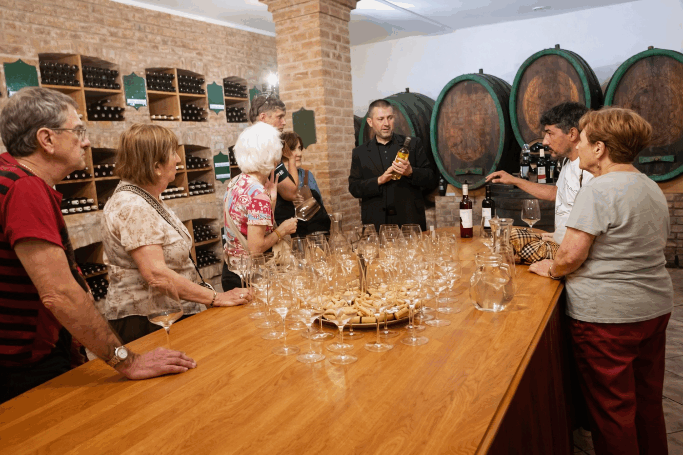 A group of visitors gathered around a long wooden table set with rows of wine glasses for a tasting session inside a traditional Slovak wine cellar, with large oak barrels and bottle-lined brick shelving in the background. A host presents a bottle of white wine, with rosé and red wines also visible on the table.