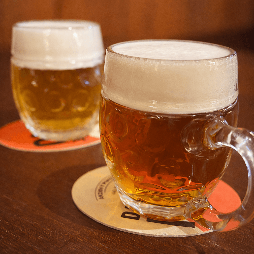 Two classic glass beer mugs filled with golden Slovak lager, topped with thick creamy foam heads, resting on branded coasters on a dark wooden pub table in Bratislava.